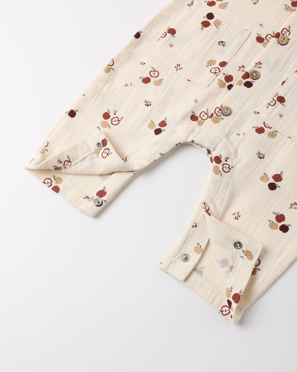 A close-up of a cream-colored cotton gauze baby romper or jumpsuit with a small red, brown, and beige fruit print, featuring a buttoned cuffed sleeve and a chest pocket.