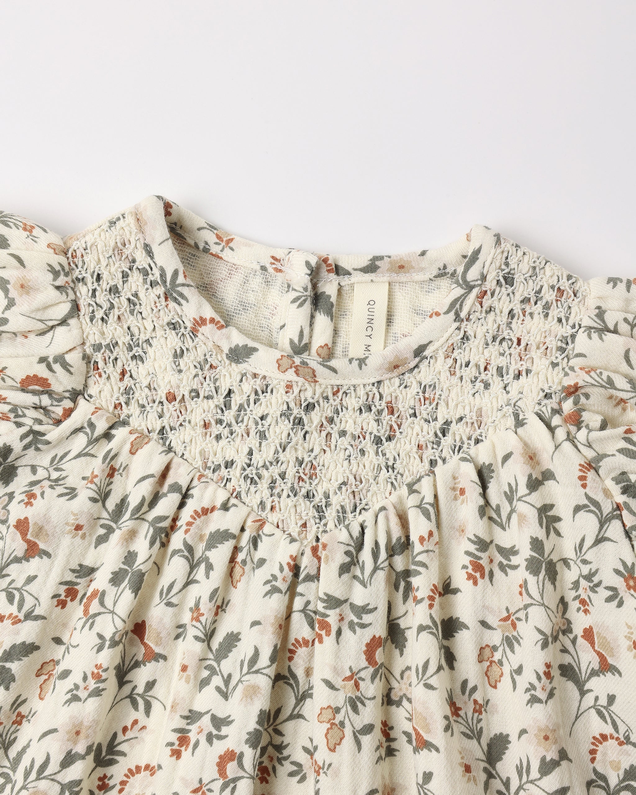 Carina Dress - Holiday Floral by Quincy Mae shown in a close-up highlighting the natural fabric, festive floral print, short bubble sleeves, and diamond smocking around the neckline.