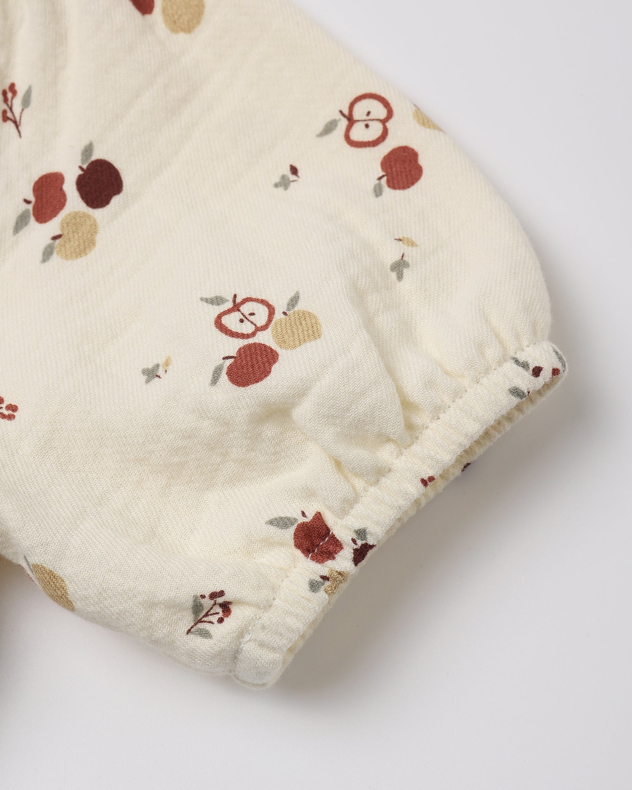 Close-up of a cream-colored fabric sleeve or cuff with a small apple and leaf print, finished with a gathered elastic hem, on a light gray background.