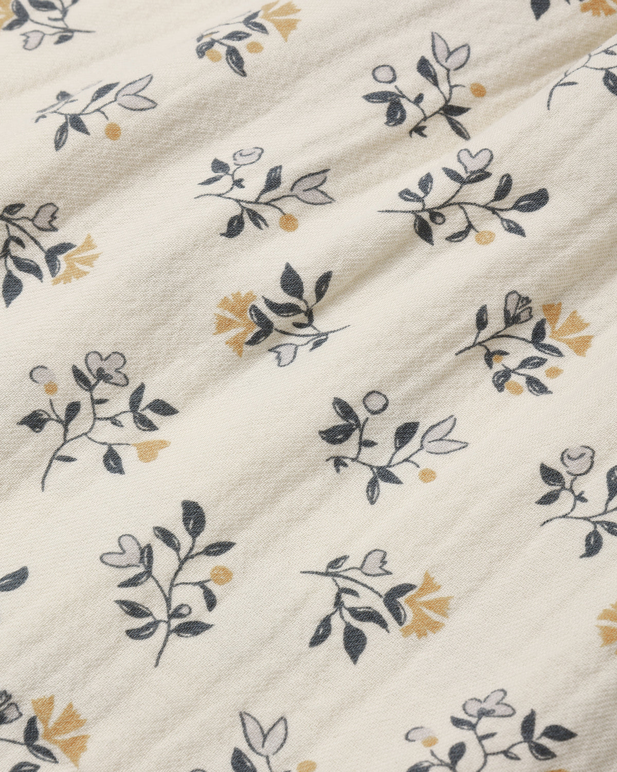 Close-up of a cream-colored fabric with a delicate botanical print featuring navy blue leaves, light gray outlines, and small mustard yellow floral accents.