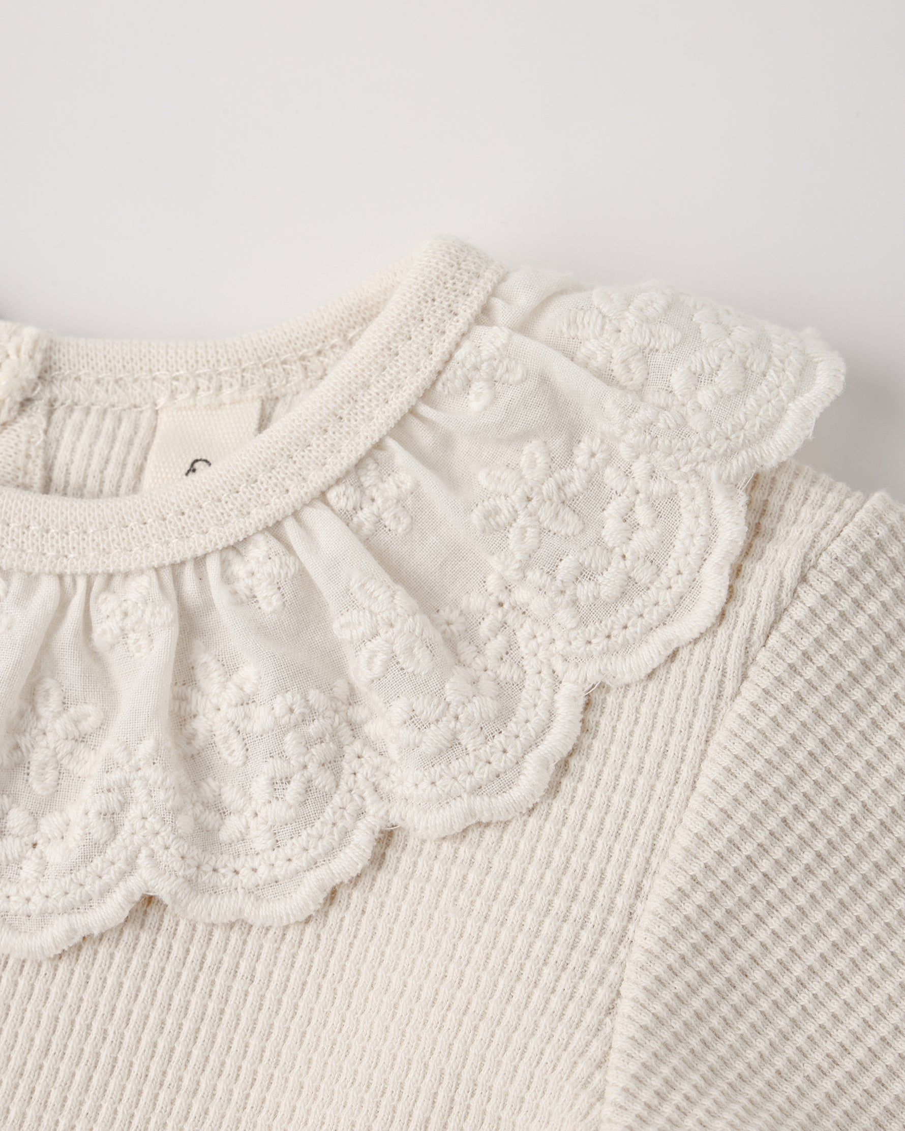 Close-up of a natural beige baby bodysuit featuring a delicate, lace-trimmed ruffled collar and textured waffle-knit fabric.
