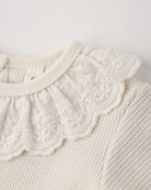 Close-up of a natural beige baby bodysuit featuring a delicate, lace-trimmed ruffled collar and textured waffle-knit fabric.