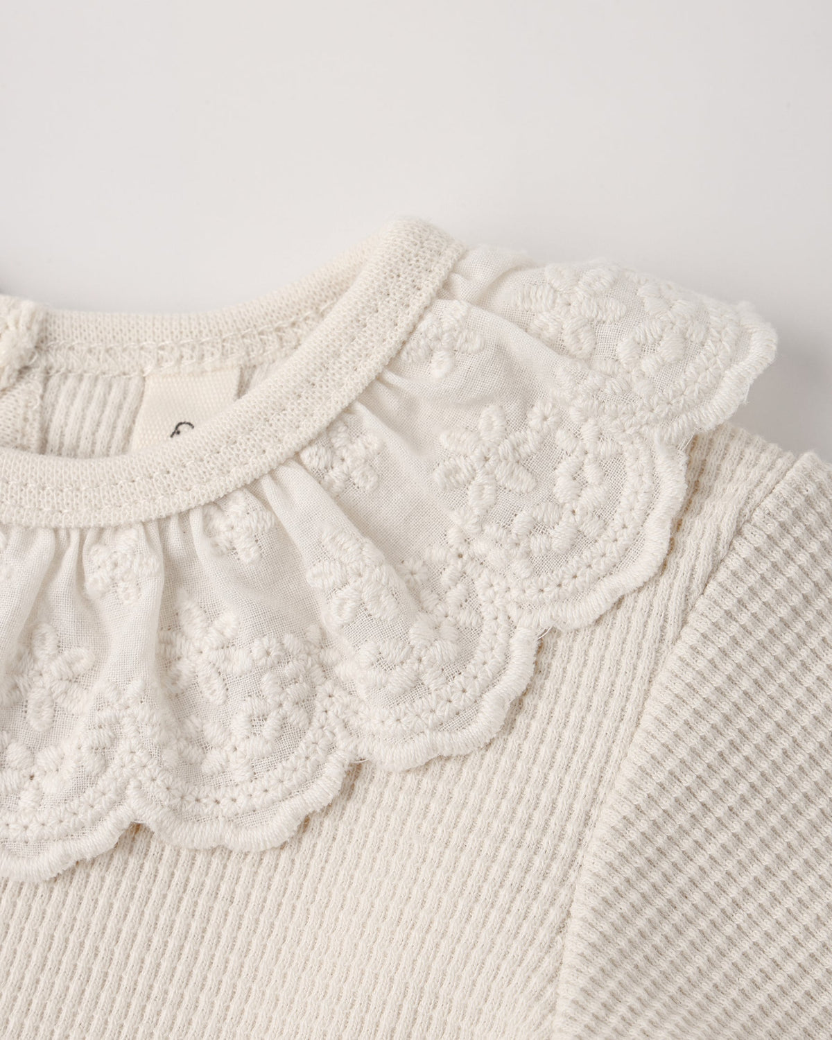 Close-up of a natural beige baby bodysuit featuring a delicate, lace-trimmed ruffled collar and textured waffle-knit fabric.