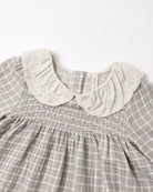 Close-up of a children's dress with a gray and white gingham pattern, featuring a smocked bodice and a large cream-colored ruffled lace Peter Pan collar.