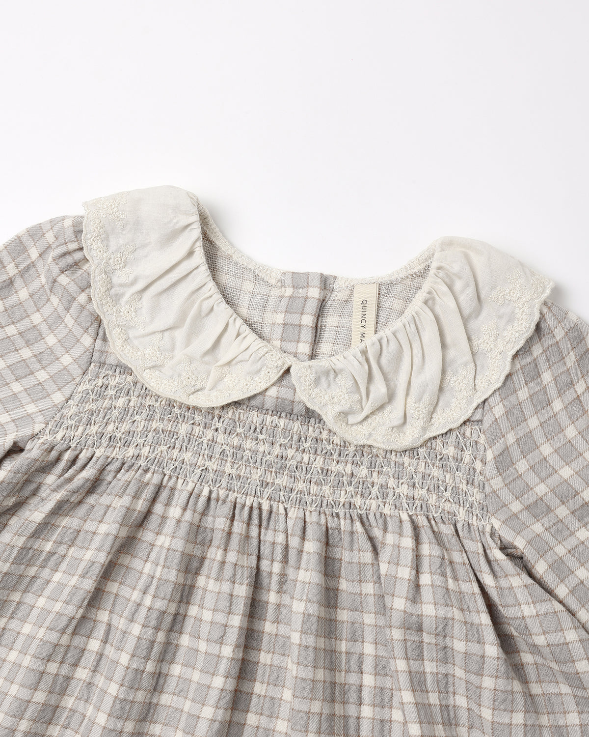 Close-up of a children's dress with a gray and white gingham pattern, featuring a smocked bodice and a large cream-colored ruffled lace Peter Pan collar.