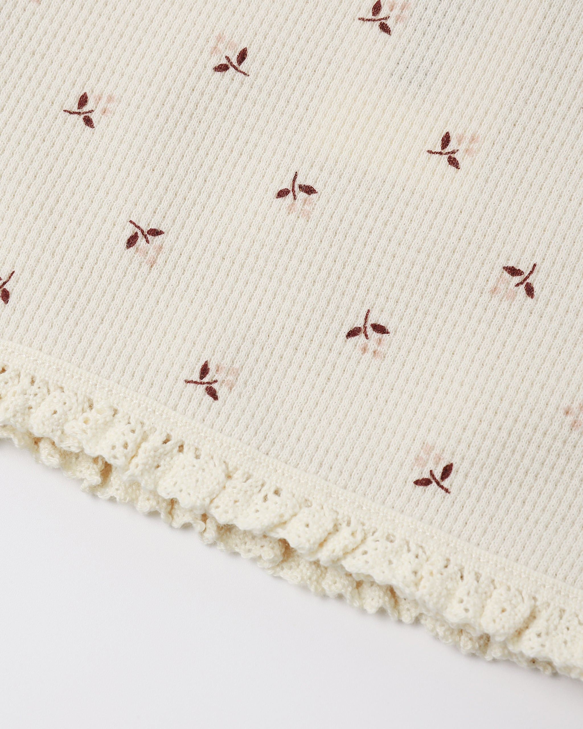 Close-up of a cream-colored ribbed knit fabric with a delicate ruffled lace trim and small embroidered floral motifs in brown and soft pink.