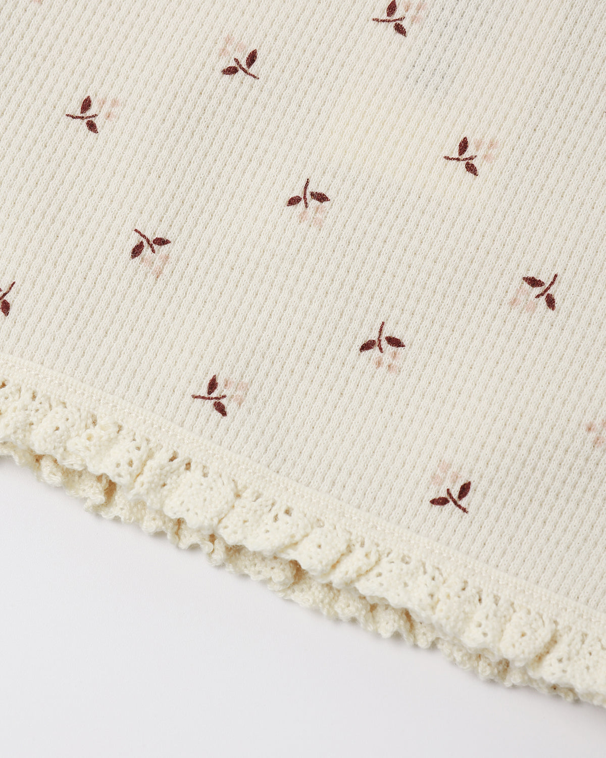 Close-up of a cream-colored ribbed knit fabric with a delicate ruffled lace trim and small embroidered floral motifs in brown and soft pink.