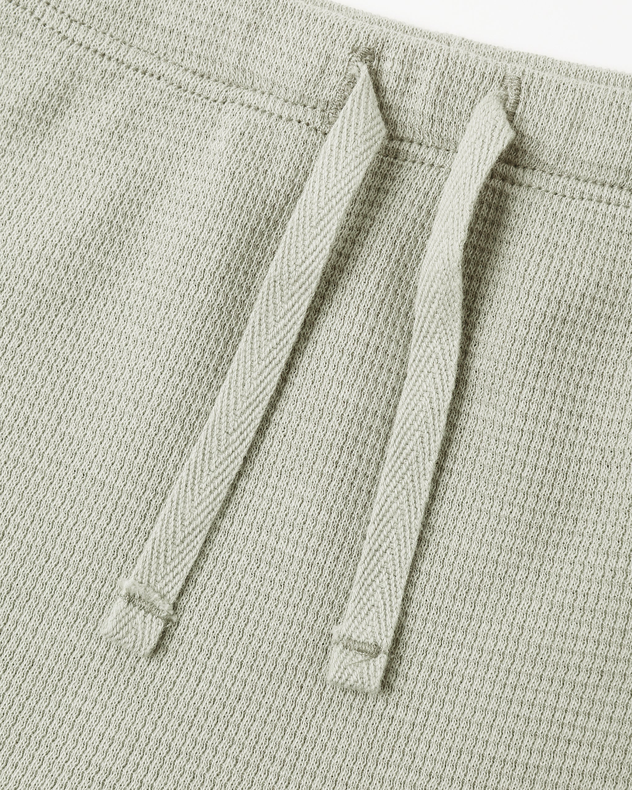 Close-up of a light sage green textured knit garment, showing two matching drawstrings against a white background.
