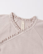 Close-up of a knit sweater in a light warm beige or taupe color, showing a waffle-knit texture, a ruffled collar, and a cream-colored interior neck label that reads 'QUINCY ART'.