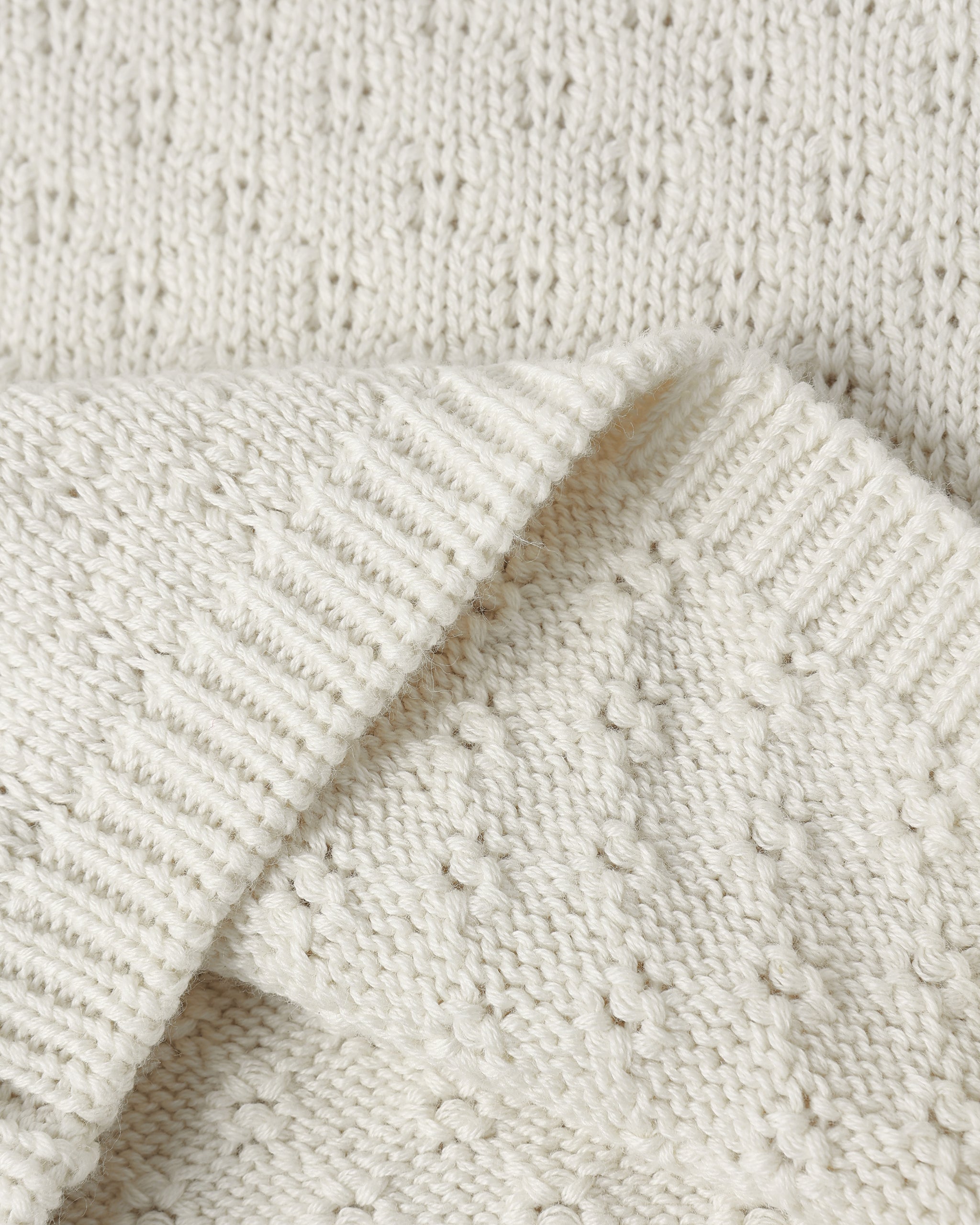 Close-up of a cream-colored knit fabric with a textured bobble pattern and ribbed trim, folded over to show the material detail.