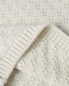Close-up of a cream-colored knit fabric with a textured bobble pattern and ribbed trim, folded over to show the material detail.
