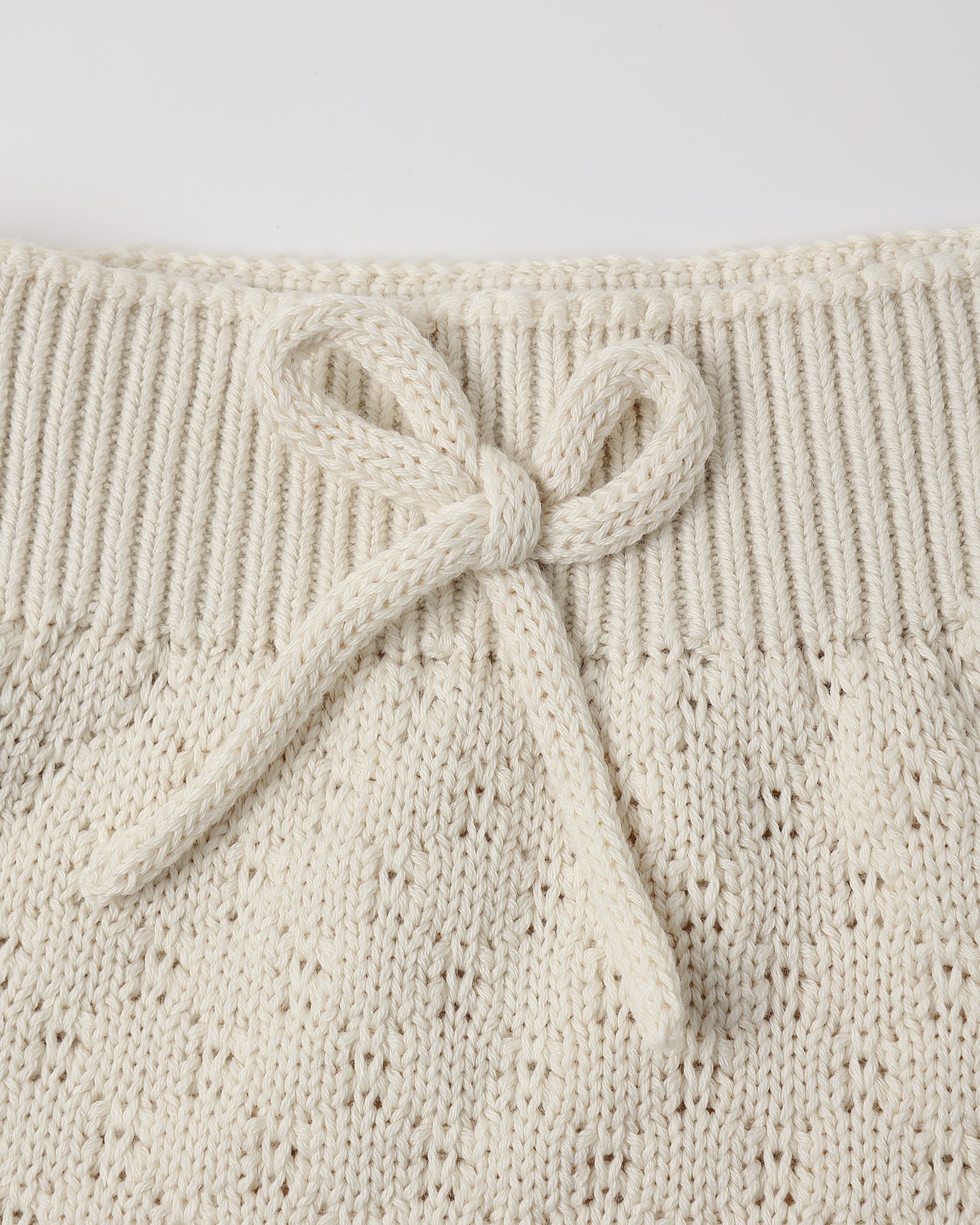 Close-up of a cream-colored knit garment featuring a ribbed waistband and a knotted bow detail, showing the textured openwork knit pattern.