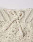 Close-up of a cream-colored knit garment featuring a ribbed waistband and a knotted bow detail, showing the textured openwork knit pattern.