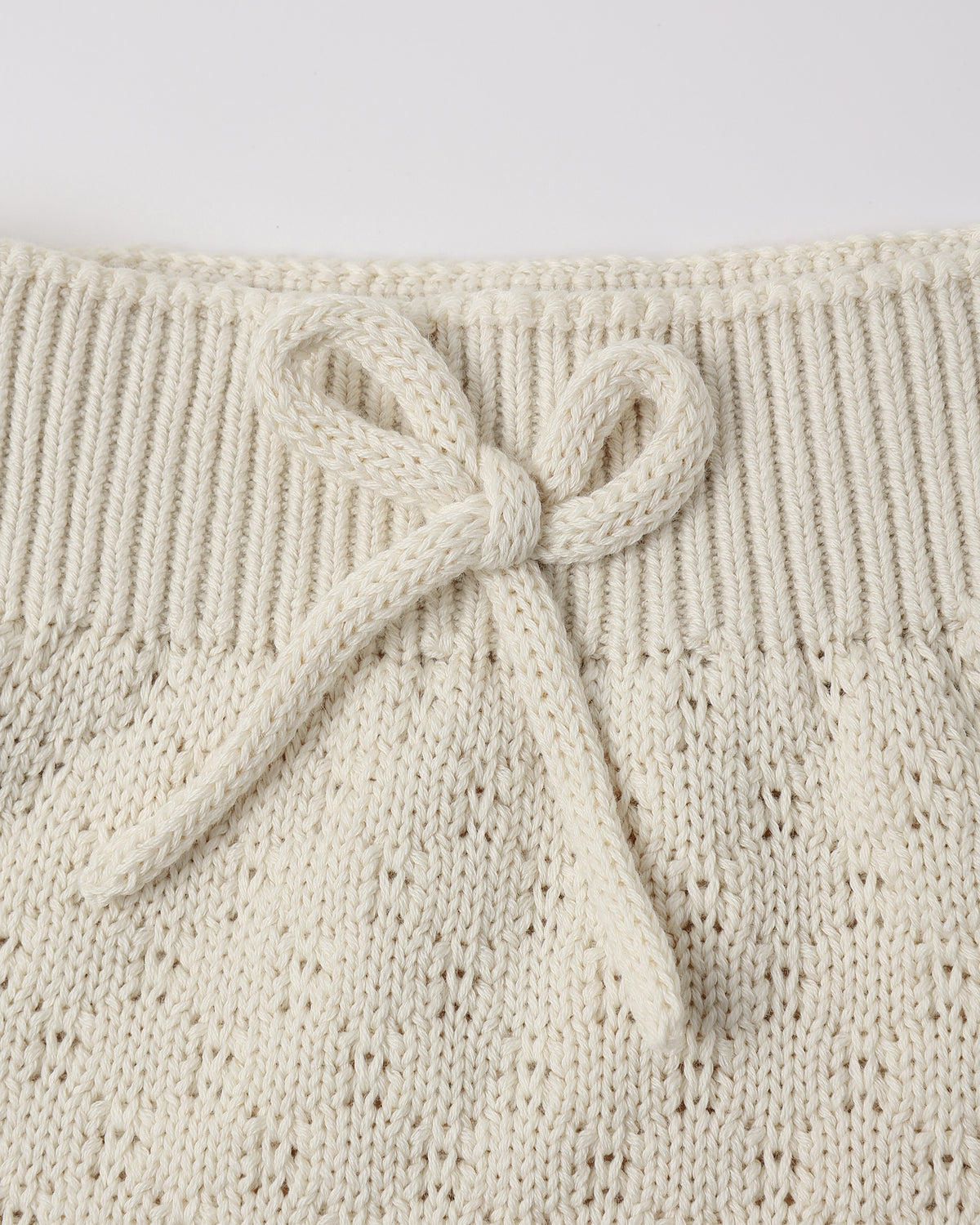 Close-up of a cream-colored knit garment featuring a ribbed waistband and a knotted bow detail, showing the textured openwork knit pattern.