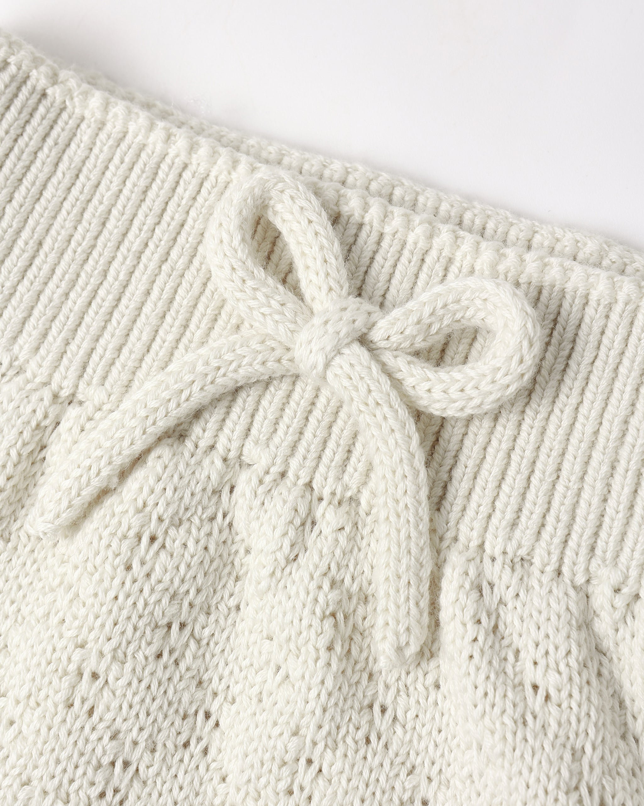Close-up of a cream-colored knit garment featuring a textured cable-knit pattern and a matching knotted bow detail at the waistband.