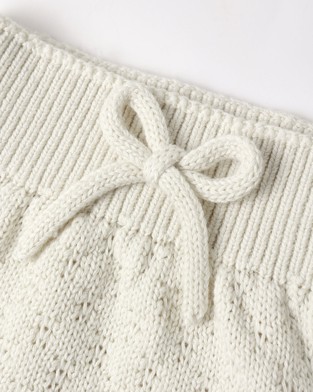 Close-up of a cream-colored knit garment featuring a textured cable-knit pattern and a matching knotted bow detail at the waistband.