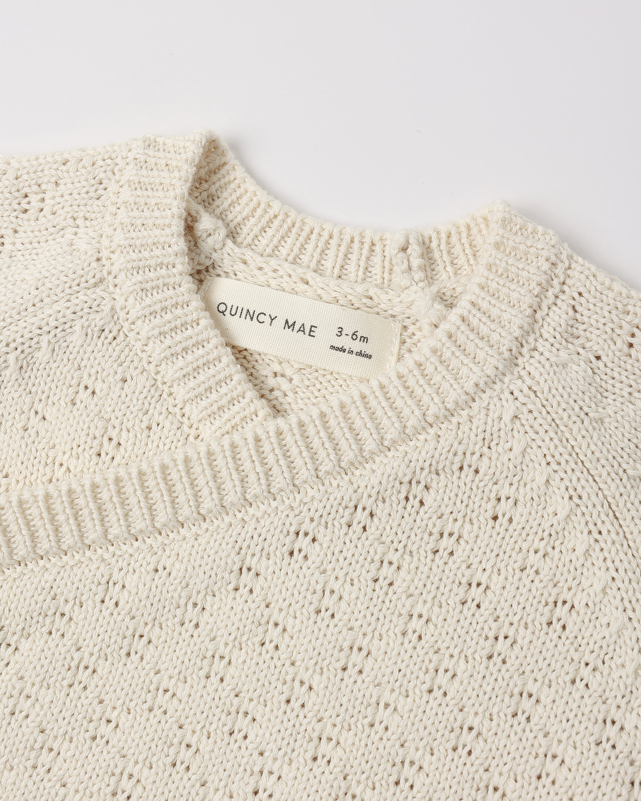 Close-up of a cream-colored knit sweater with a visible neck label reading 'QUINCY MAE 3-6m made in Spain'.
