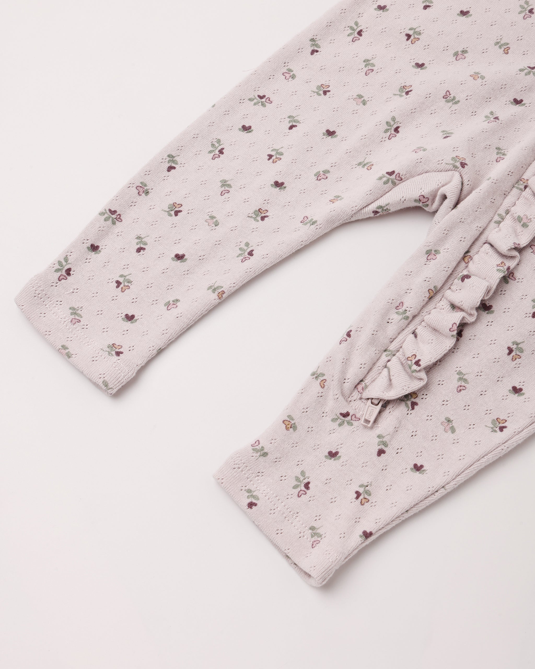 Close-up of a baby or toddler long-sleeve romper or pajama top in a pale blush pink eyelet fabric with a small scattered floral print, featuring a ruffled trim detail on the side.
