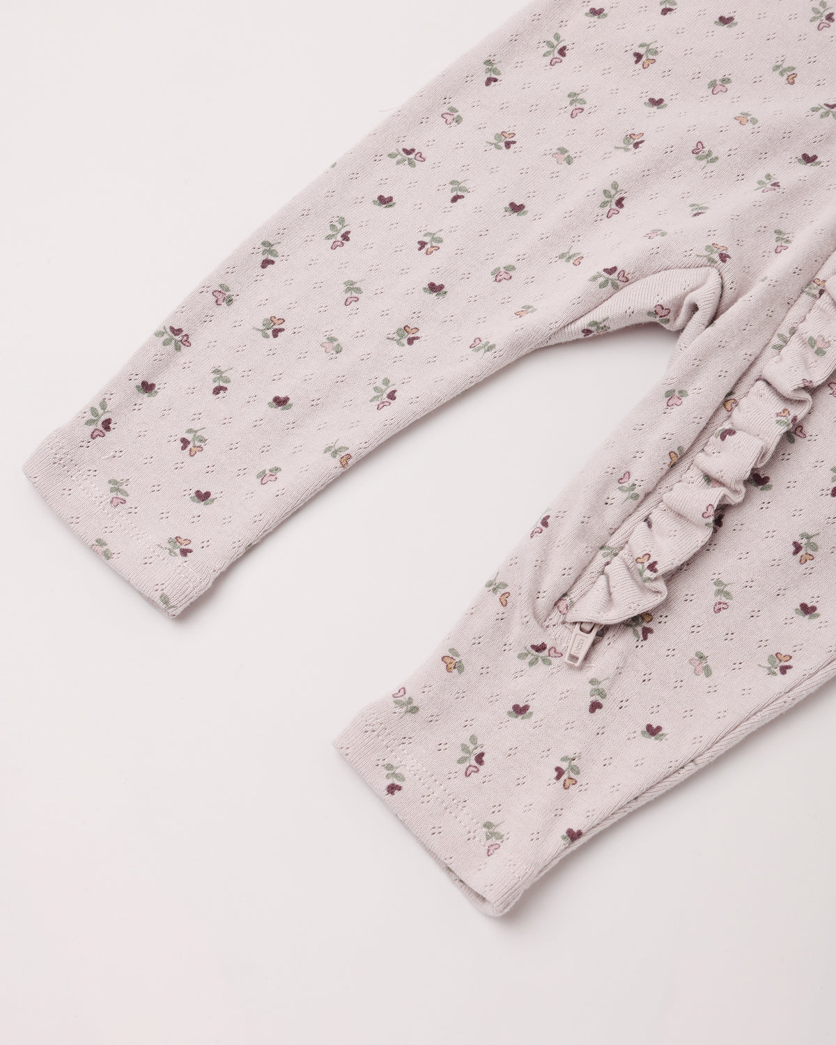 Close-up of a baby or toddler long-sleeve romper or pajama top in a pale blush pink eyelet fabric with a small scattered floral print, featuring a ruffled trim detail on the side.