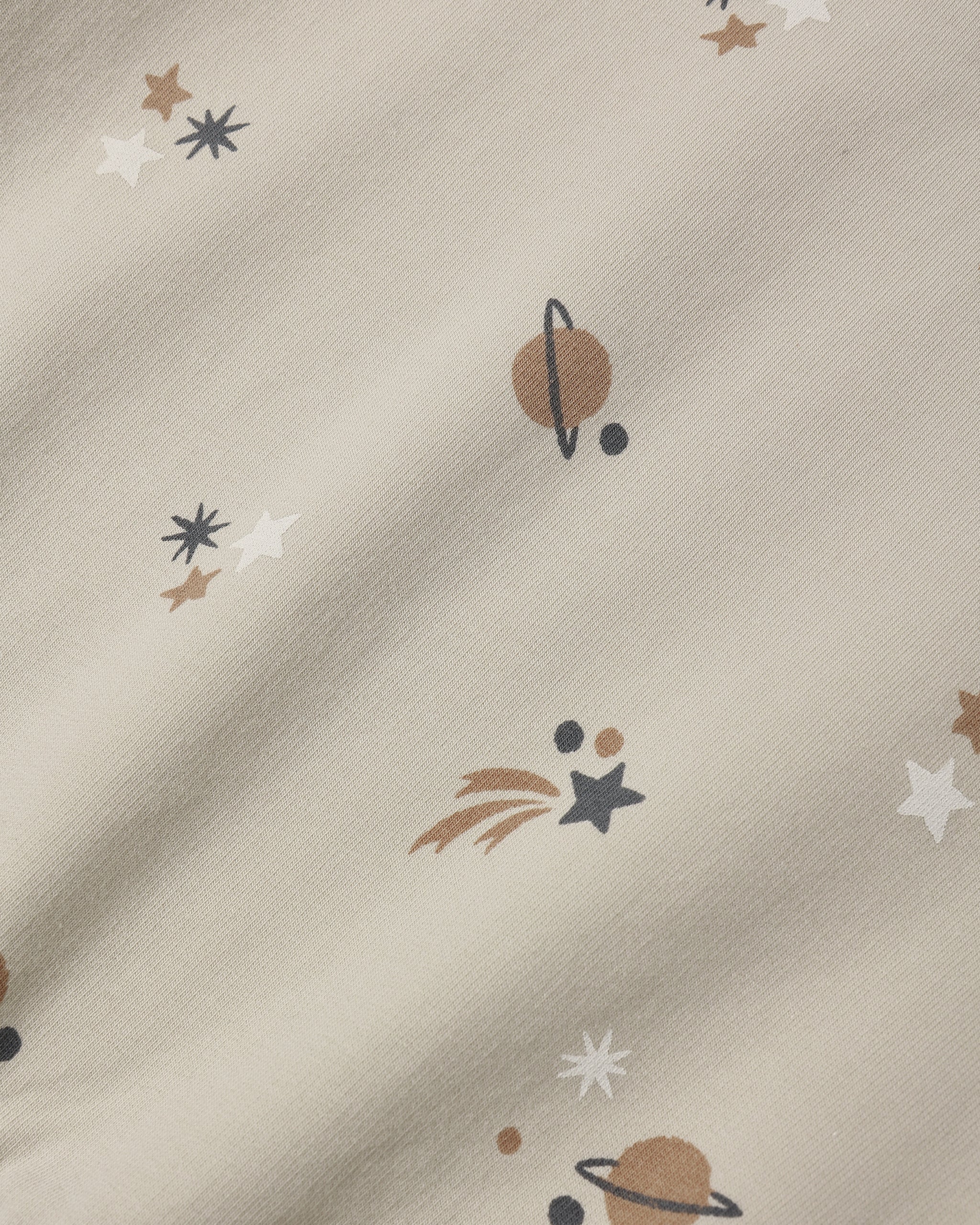 A close-up of a cream-colored fabric with a printed space theme, featuring planets, stars, and shooting stars in brown, white, and dark gray.
