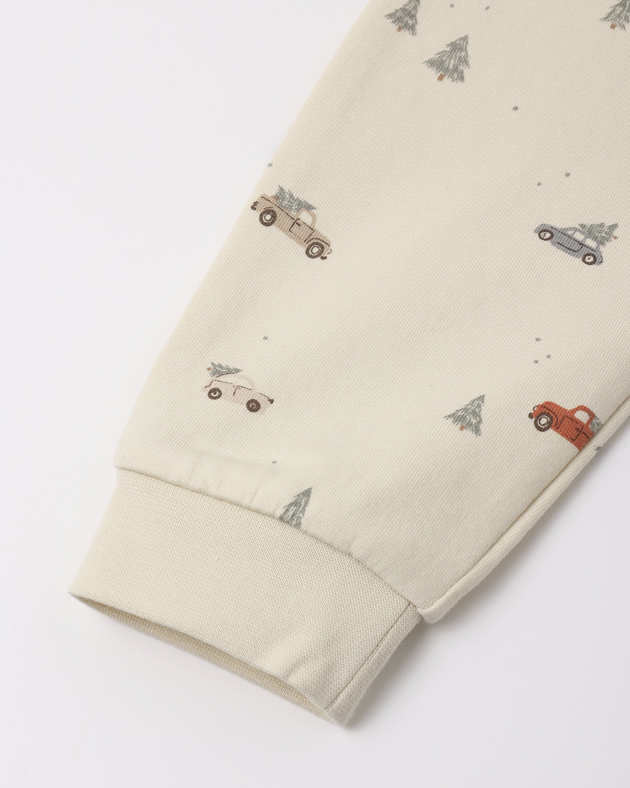 Close-up of cream-colored organic cotton fabric featuring a "Holiday Drive" print with tiny vintage cars and pine trees, showing a ribbed sleeve cuff.