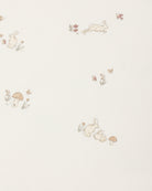 A close-up of a cream-colored fabric or wallpaper featuring a delicate, repeating pattern of small rabbits, mushrooms, flowers, and butterflies in soft beige, muted brown, and pale green tones.