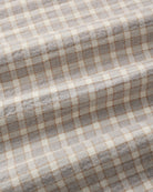 A close-up of a woven fabric with a soft plaid pattern in light gray, cream, and thin tan lines, showing the texture and folds of the material.