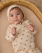 An infant wearing the Quincy Mae red bows print long-sleeve top and matching bonnet, lying comfortably in a woven basket.