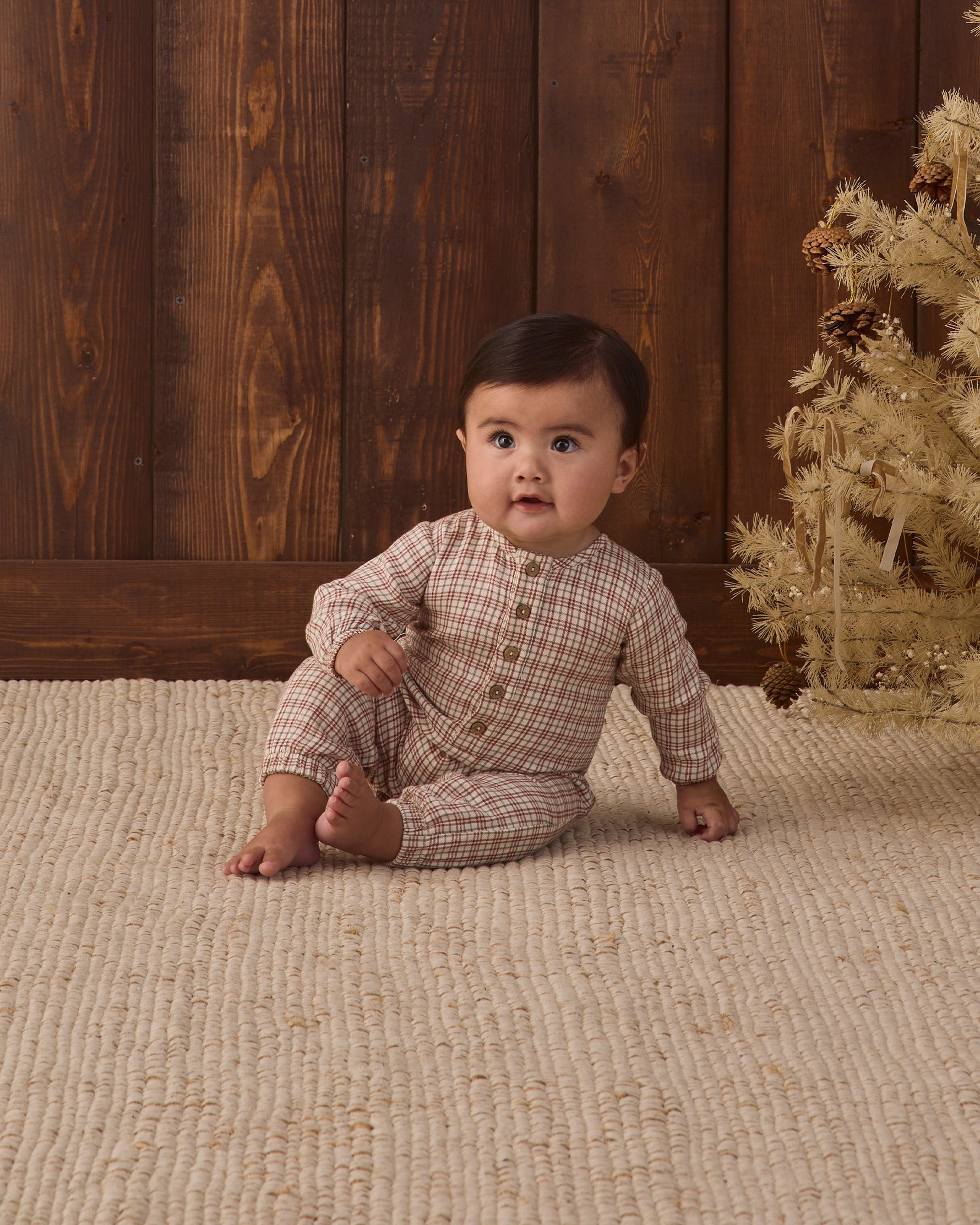 A baby crawling on a textured cream rug in front of a wood-paneled wall, wearing a long-sleeve Christmas plaid jumpsuit.