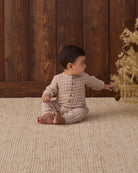 A baby sitting on a textured cream rug in front of a wood-paneled wall, wearing a long-sleeve Christmas plaid jumpsuit with a button placket on the chest.