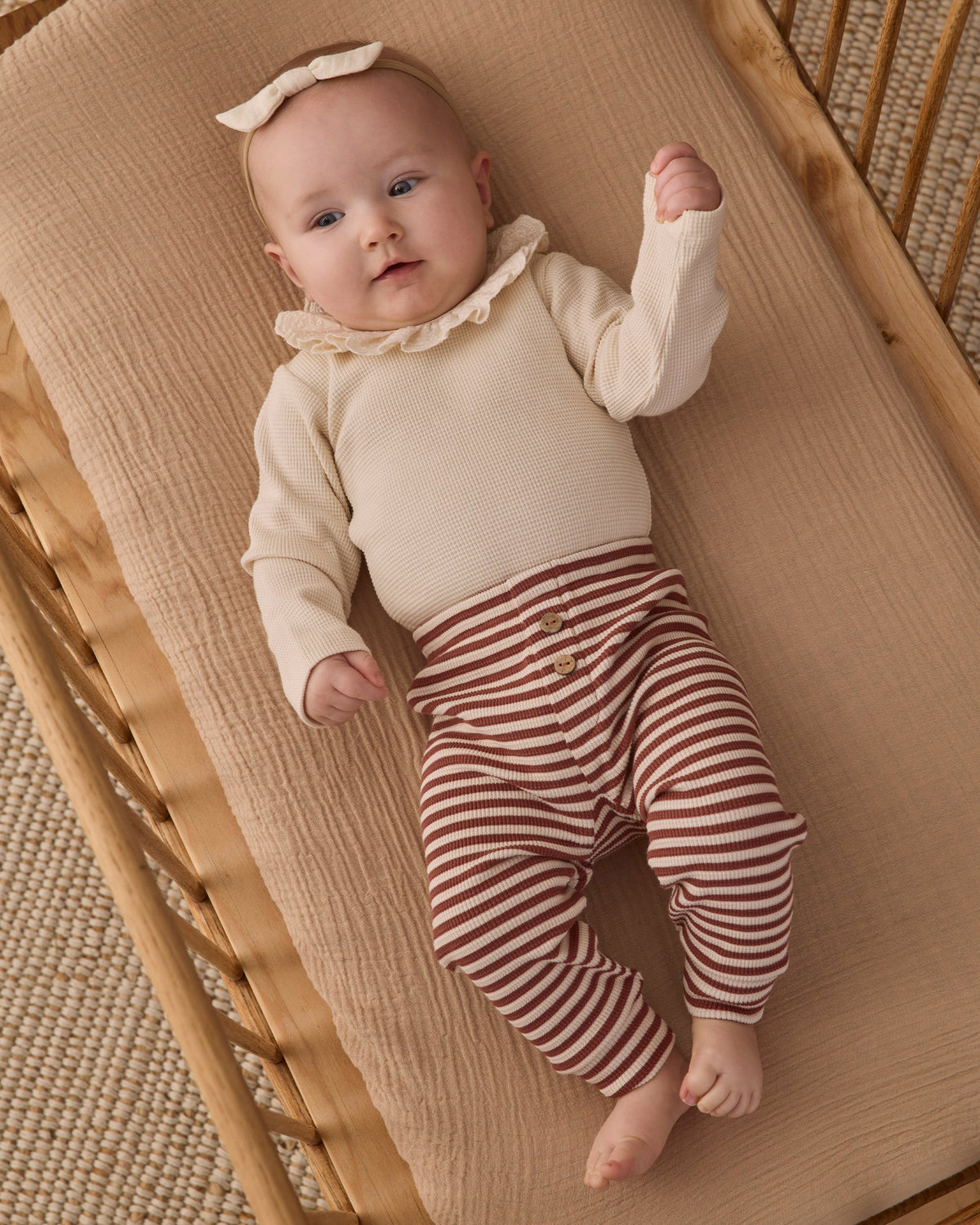 A baby lying on a woven bassinet, wearing a long-sleeve natural beige ruffled collar bodysuit and brown-and-white striped pants, wearing a small hair bow.