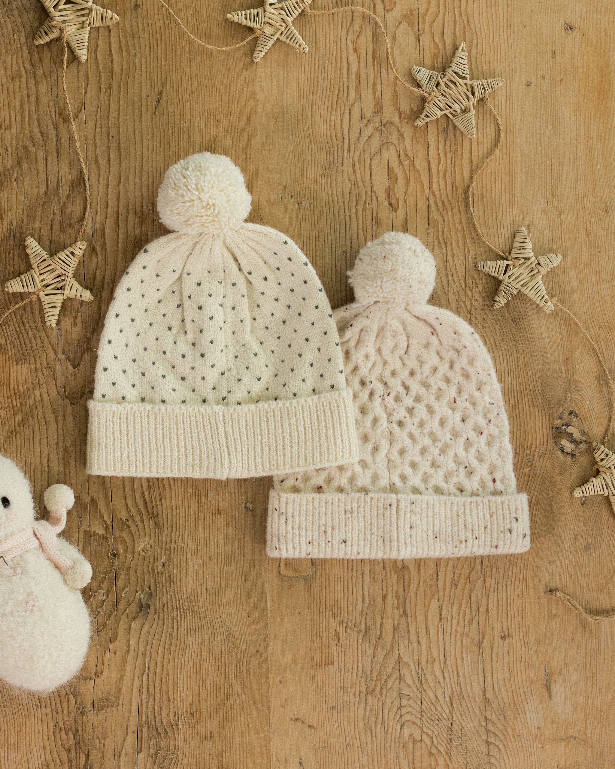 Two cream-colored knit baby hats with pom-poms on a wooden surface with decorative stars.