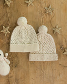 Two cream-colored knit baby hats with pom-poms on a wooden surface with decorative stars.