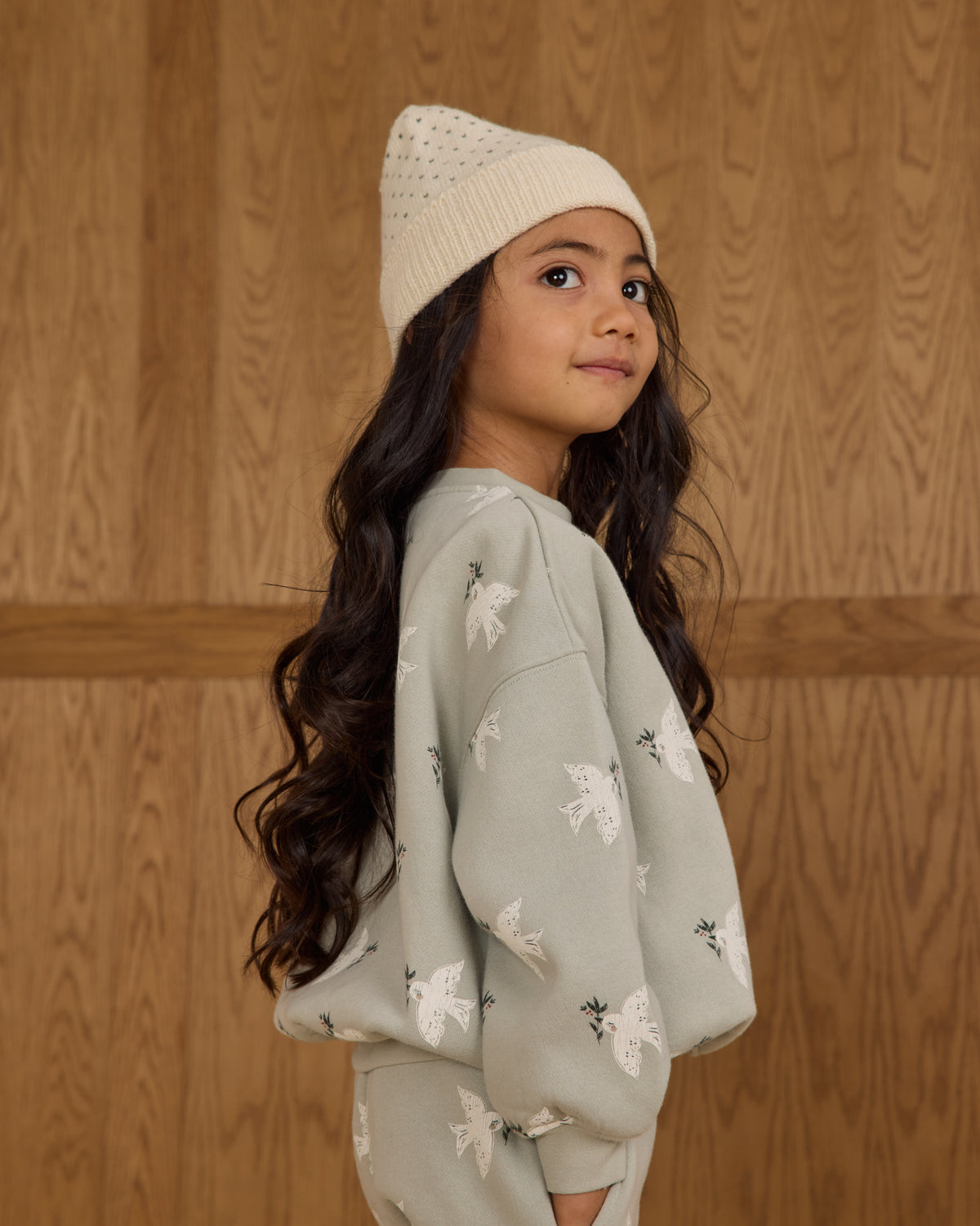 Young girl wearing a light gray floral sweater and cream beanie against a wooden background