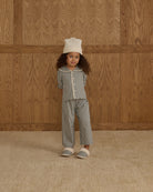 Child wearing a checkered outfit with a white hat against a wooden paneled wall.