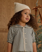 Young girl wearing a checkered dress and knit hat against a wooden background