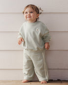 A toddler wearing a sage-green relaxed sweatshirt and matching sweatpants set with side-stripe detailing from Rylee + Cru.