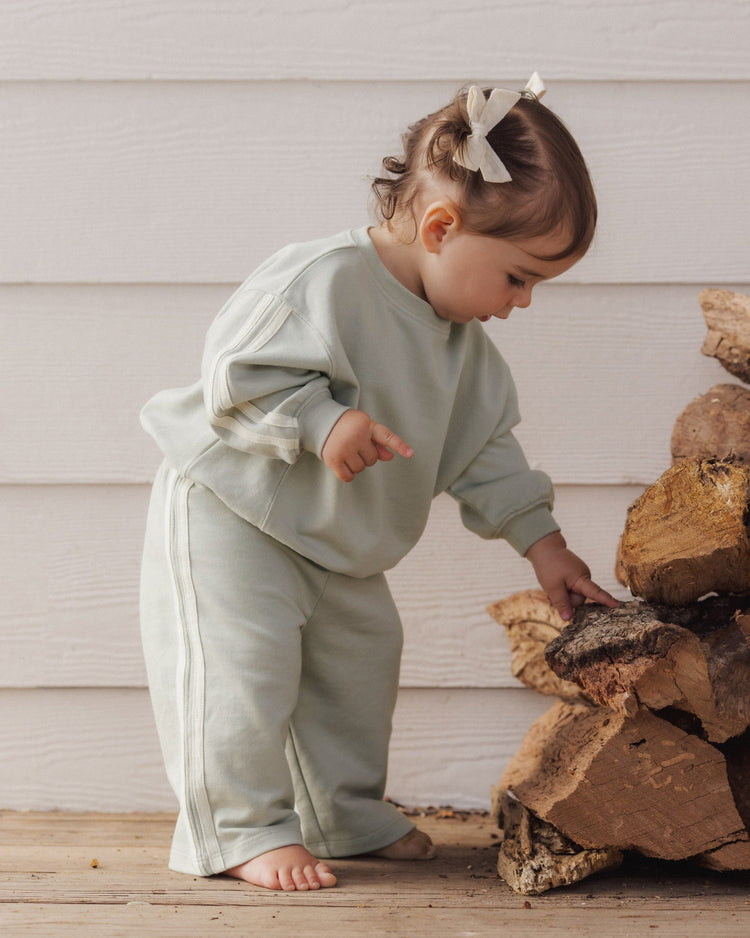 A toddler wearing a sage-green relaxed sweatshirt and matching sweatpants set with side-stripe detailing from Rylee + Cru.