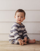 A baby sitting on a wooden floor in front of a white wood-paneled wall, wearing a long-sleeve striped one-piece pajama in navy blue and cream, holding a small wooden toy.