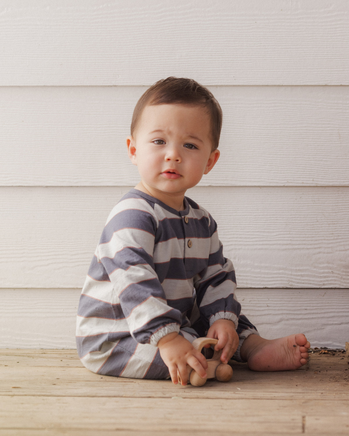 A baby sitting on a wooden floor in front of a white wood-paneled wall, wearing a long-sleeve striped one-piece pajama in navy blue and cream, holding a small wooden toy.