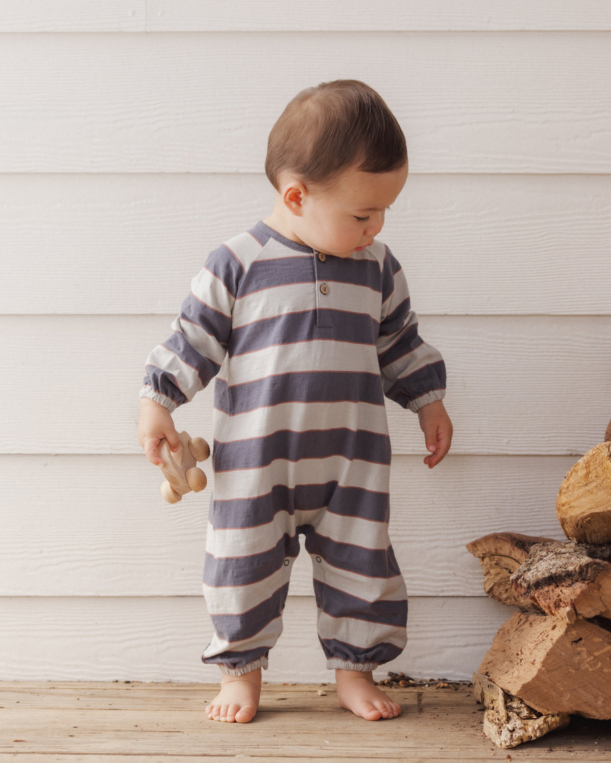 A toddler standing barefoot against a white wood-paneled wall, wearing a long-sleeve footless one-piece jumpsuit with horizontal navy and off-white stripes. The child is holding a small wooden toy.