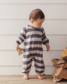 A toddler standing barefoot against a white wood-paneled wall, wearing a long-sleeve footless one-piece jumpsuit with horizontal navy and off-white stripes. The child is holding a small wooden toy.