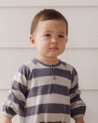 A toddler boy wearing a long-sleeve henley shirt with horizontal navy and beige stripes, standing against a white wood-paneled wall.
