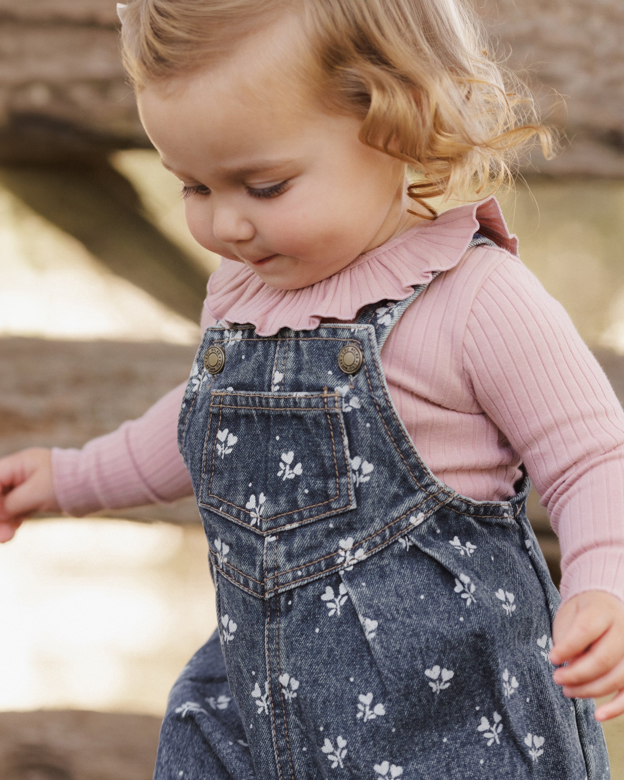 A toddler girl wearing a dusty pink ribbed long-sleeve top and dark blue denim overall with a white floral pattern, looking down in an outdoor setting.