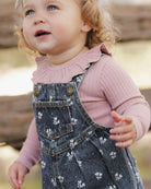 A toddler girl wearing a dusty pink ribbed long-sleeve top with a ruffled collar and dark blue denim overall dress with a white floral print, photographed outdoors.
