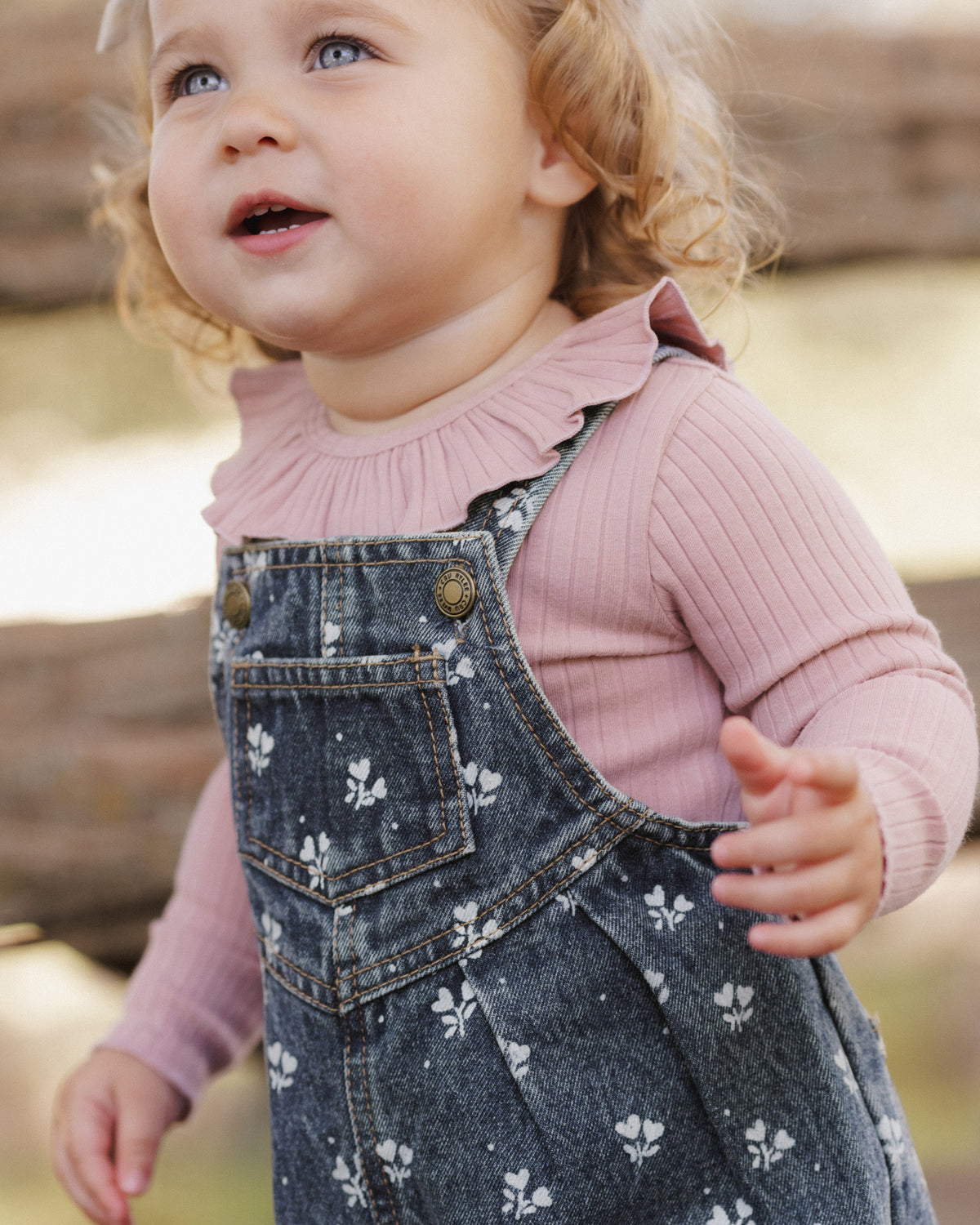 A toddler girl wearing a dusty pink ribbed long-sleeve top with a ruffled collar and dark blue denim overall dress with a white floral print, photographed outdoors.