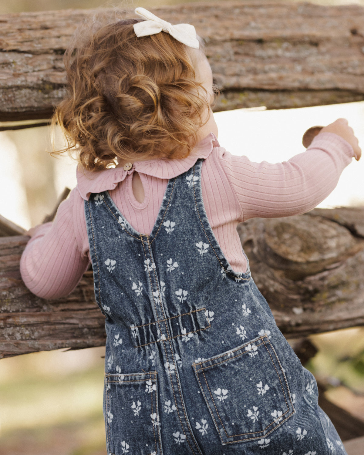 A toddler seen from behind wearing a pink ribbed long-sleeve top and a blue denim overall with a white floral print, leaning on a rustic wooden fence.