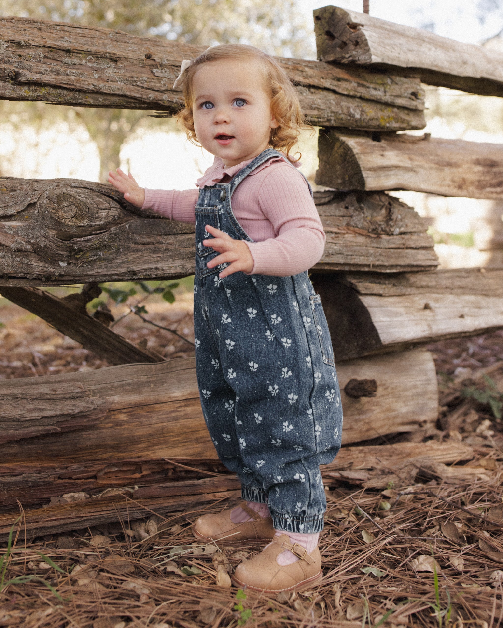 A toddler girl wearing a pink long-sleeve top and dark blue patterned overall-style romper stands outdoors in front of a rustic wooden fence on a wooded path.