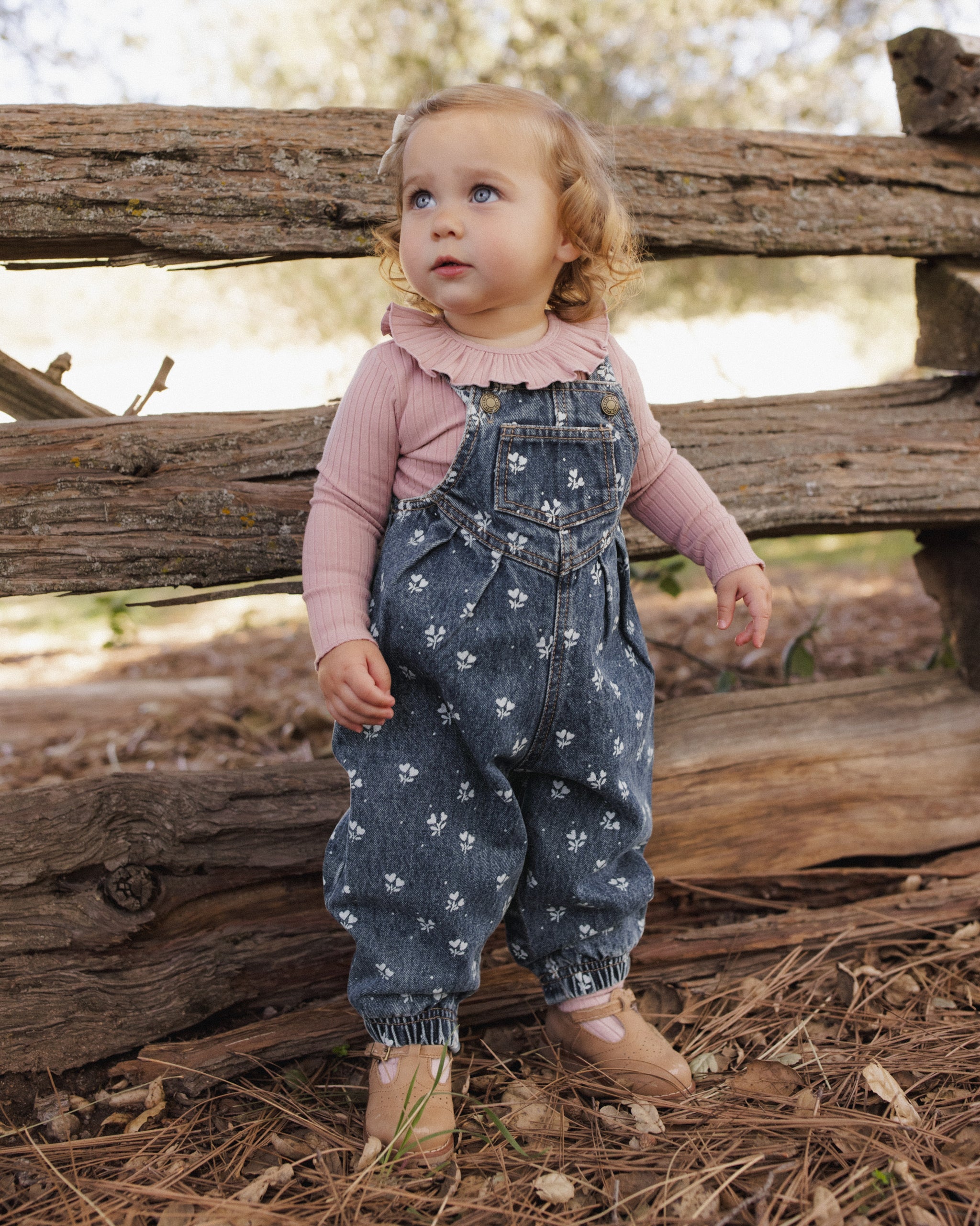 A toddler standing outdoors in front of a rustic wooden fence, wearing blue denim overall with a white floral print over a dusty pink long-sleeve ruffled shirt and tan shoes.