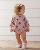 A toddler wearing a mauve-pink long-sleeve crewneck bubble romper featuring an all-over brown daisy floral print by Rylee + Cru.